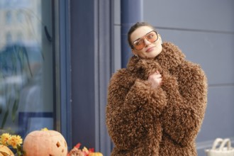 A young woman stands outside a shop, dressed in a fluffy brown coat and stylish sunglasses.