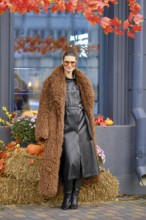 A stylish woman poses outside a farmer's grocery shop decorated for autumn, wearing a brown fur
