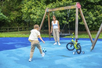 Young boy kicking a soccer ball on a playground while mother watches, with a swing set and bicycle