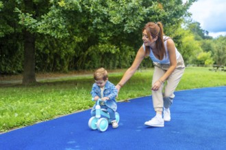 Young mother helping her son learn to ride a balance bike on a blue surface in a green park,