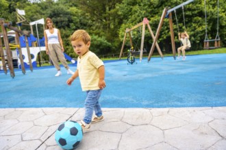 Happy toddler kicking a vibrant soccer ball at the playground, with mother and sister enjoying