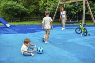 Two brothers are playing with a ball and a bicycle at the playground while their mother watches
