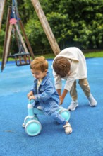 Older brother is helping his younger brother to ride a balance bike on a blue playground surface