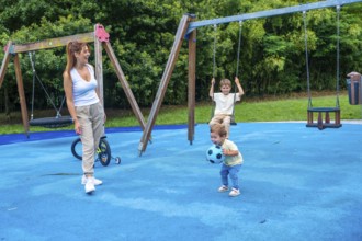 Mother enjoying a sunny day at the playground, watching her children happily playing with a ball