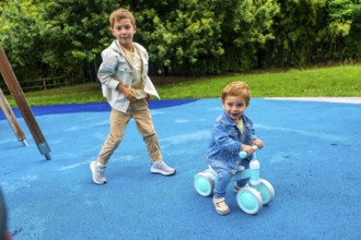 Two brothers are spending time together on a playground, one riding a balance bike and the other
