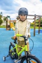 Happy boy wearing helmet learning to ride a bicycle with training wheels in a playground on a sunny