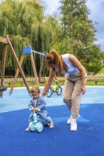 Young mother helping her toddler son learn to ride a balance bike on a playground's soft blue