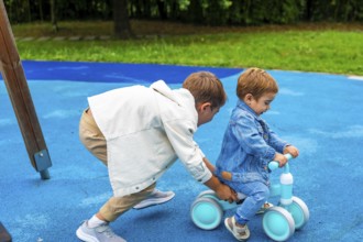 In a vibrant playground setting, an older brother supports his younger sibling as he learns to ride