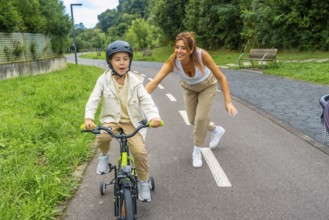 Happy mother running alongside her son, guiding him as he learns to ride a bicycle with training
