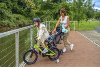 Young mother pushing a stroller with a baby inside while watching her other son riding a bicycle