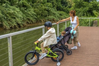 Mother walking with her baby in a stroller and her older son riding a bicycle on a wooden walkway