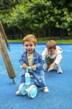 Happy toddler boy riding a balance bike on a playground with his older brother cheering him on,