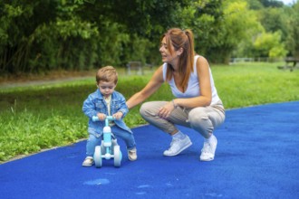 Young mother crouching next to her son helping him learn to ride a balance bike on a blue surface