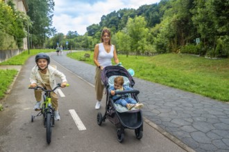 Mother pushing a stroller while walking alongside her son riding a bicycle in a vibrant park,