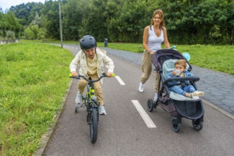 Mother walking with her baby in a stroller while older son riding his bicycle on a paved path,