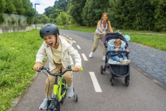 Child wearing a helmet is riding a bicycle and shouting, while his mother is pushing a stroller