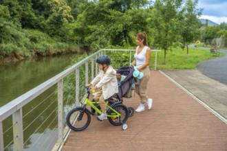 Young mother pushing a stroller with a baby inside and watching her other son riding a bicycle on a
