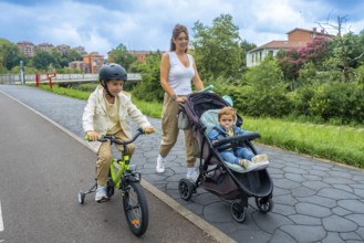 Mother enjoys a leisurely stroll in an urban park, pushing a stroller with her baby while her older