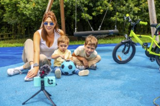 Content creator mother filming video with her sons and a soccer ball on a playground with a swing