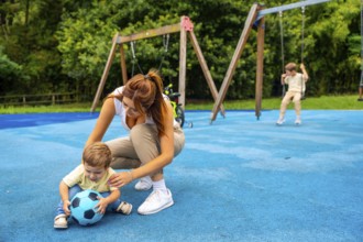 Young mother crouching next to her toddler son playing with a ball on a blue playground surface