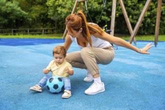 Young mother crouching beside her toddler son, guiding him as he plays with a soccer ball on a