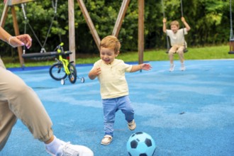 Happy toddler boy kicking soccer ball at playground with family, enjoying a playful moment on a