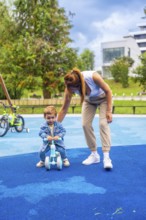 Young mother guiding her son as he learns to ride a balance bike on a playground featuring a soft