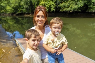 Mother and her two sons are taking a selfie on a wooden dock next to a river, surrounded by lush
