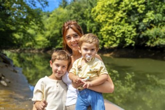Happy mother holding her baby and posing with her older son by the river in a sunny summer day,