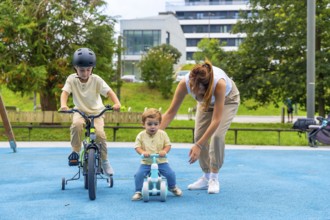 Young mother assisting two children riding bikes in a sunny playground, with the older boy on a