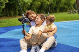 Mother and two sons are sitting on playground's rubber surface, holding a smartphone attached to a