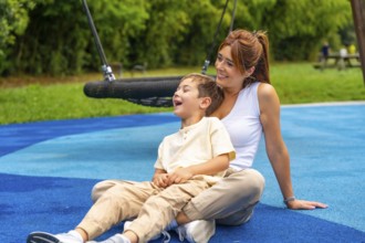Happy mother and son sitting together on blue rubber flooring near a swing, laughing joyfully and