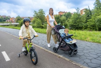 Young mother pushing a stroller with her baby boy, while her older son rides a bicycle with a