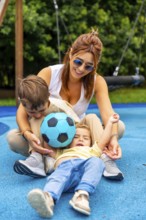 Happy mother and two children playing with a soccer ball on a playground's blue rubberized surface,