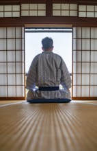Young man wearing kimono sitting in traditional Japanese living room with tatami mats and shoji