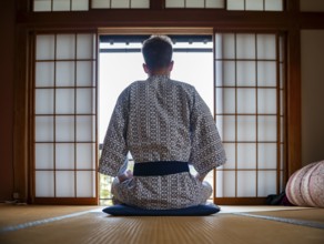 Young man wearing kimono sitting in traditional Japanese living room with tatami mats and shoji