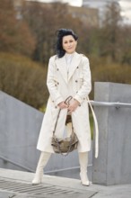 A fashionable woman stands confidently in a city park during autumn. She wears a long cream coat