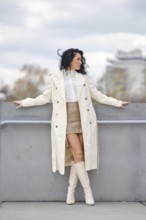 A woman showcases her autumn street style while standing on a balcony. She wears a long white coat