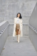 A woman strolls down a sleek walkway, wearing a beige trench coat over a white blouse and a stylish