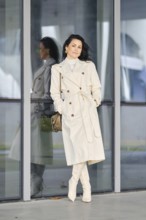 A woman showcases her autumn street style, wearing a cream coat and boots while leaning against a