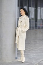 A confident woman stands next to a concrete pillar, dressed in a cream trench coat and matching
