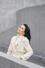 A woman stands confidently in a fashionable trench coat, gazing upward. The backdrop features sleek