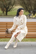 A woman dressed in a stylish cream trench coat and knee-high boots sits gracefully on a wooden