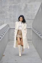 A young woman walks confidently down a sleek concrete staircase, wearing a light-colored trench