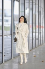 A stylish woman strolls along a sleek urban walkway. She wears a light trench coat and matching