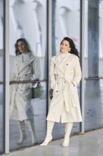 Dressed in a stylish white trench coat and matching boots, a woman stands confidently against a