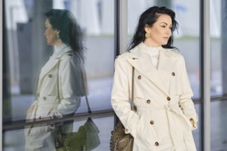 A woman dressed in a fashionable trench coat stands outside, her reflection visible in the glass