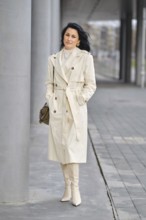 A confident woman strolls along a modern urban walkway, dressed in a light trench coat and matching