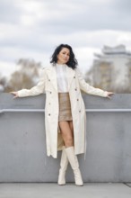 A woman stands confidently against a gray barrier, wearing a stylish long coat, knee-high boots,