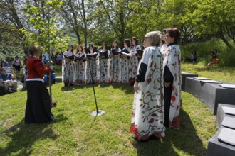 Women's choir in white colorful robes singing outdoors, surrounded by trees and audience, choir at